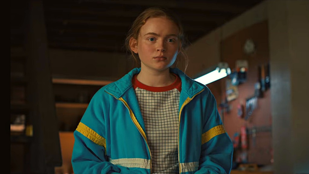 Max Mayfield en veste bleue à bandes jaunes, col rouge, dans un atelier