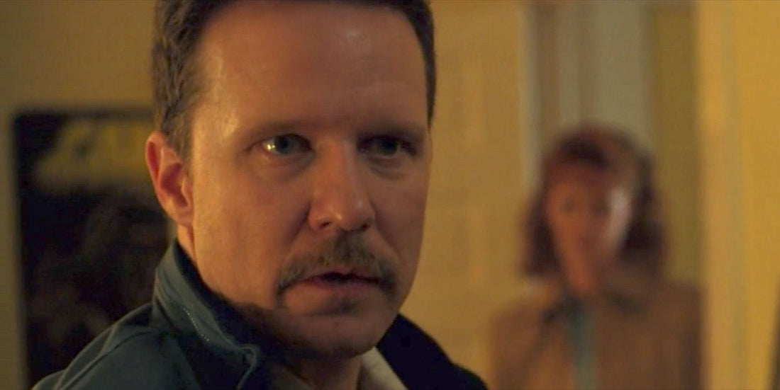 Neil, moustache et veste sombre, dans le couloir, Susan floue derrière lui.