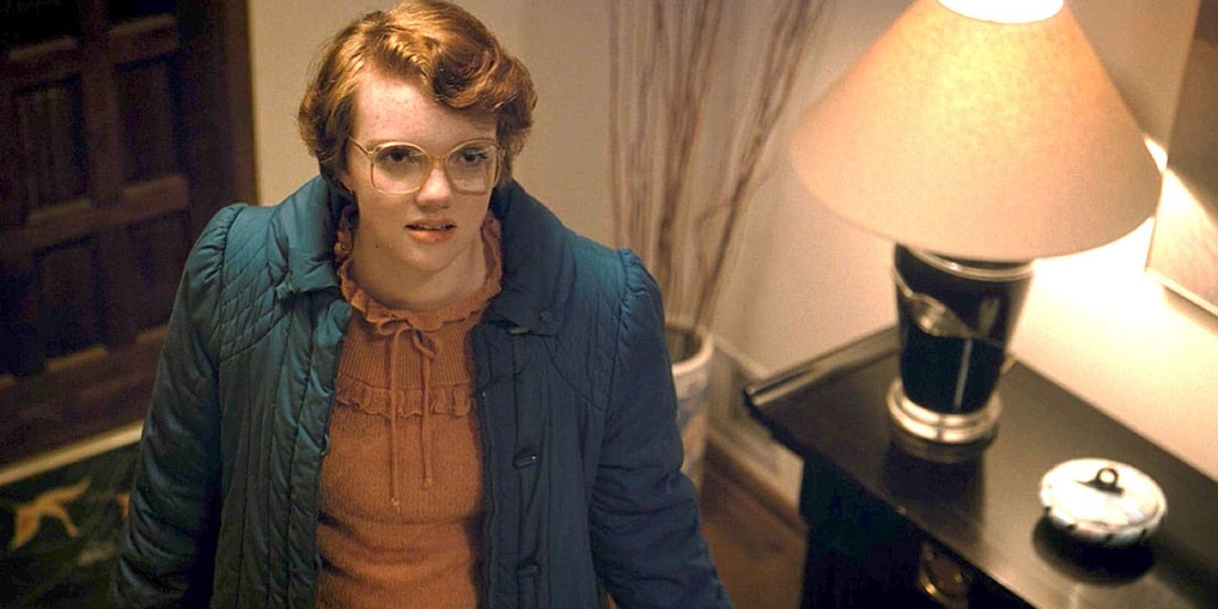 Shannon Purser en Barbara Holland, lunettes et manteau bleu, dans une entrée.